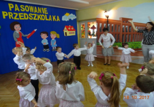 Odświętnie ubrane dzieci tańczą z rękoma w górze na tle dekoracji. Dwie nauczycielki tańczą z dziećmi.